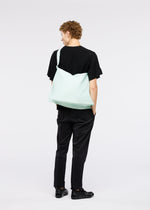 A person with short curly hair dressed in black, facing away with hands in pockets, carries the large pale green ROLL bag by GOOD GOODS ISSEY MIYAKE over their shoulder. The background is plain white.