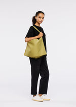 A woman stands against a white background, wearing black t-shirt and pants with white sneakers. She carries the elegant ROLL shoulder bag in olive green from GOOD GOODS ISSEY MIYAKE, looking calmly at the camera.
