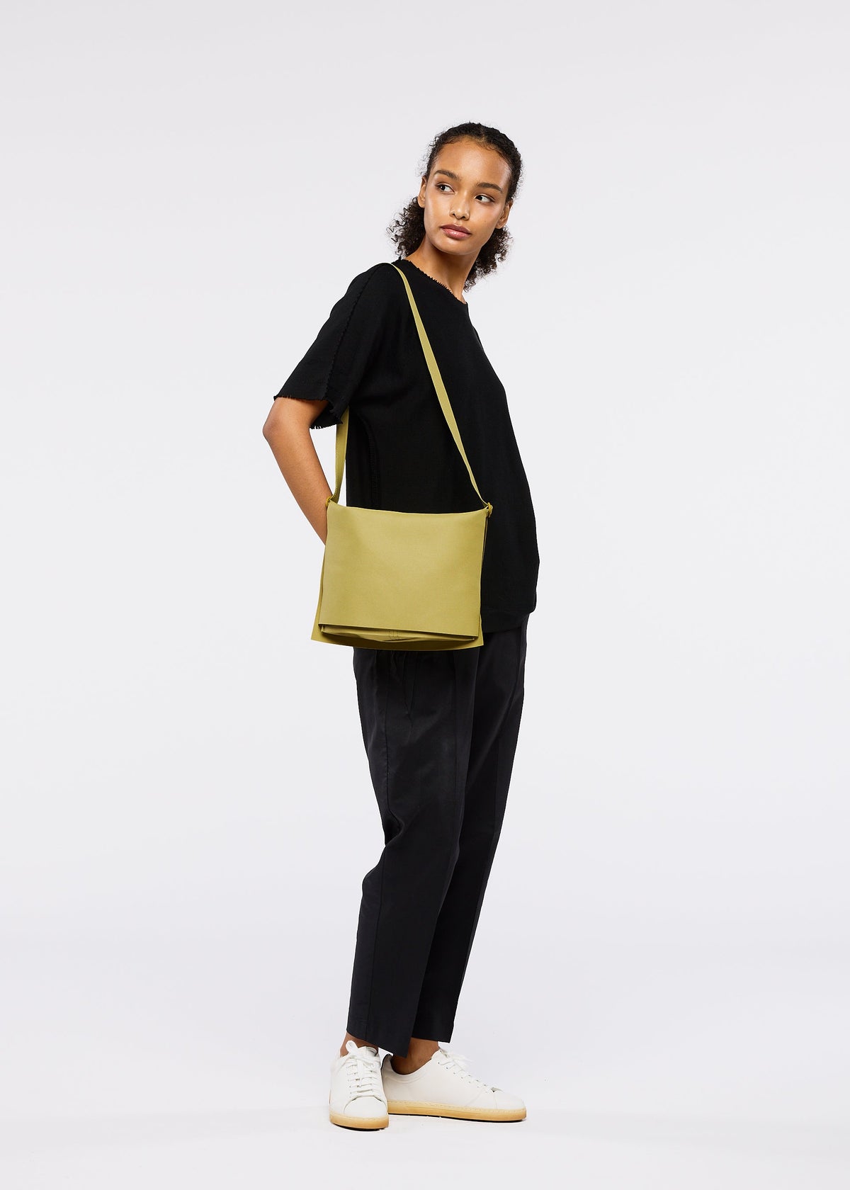 A person stands against a plain white background, wearing a black t-shirt, black pants, white sneakers, and carrying the ROLL lightweight shoulder bag in light green by GOOD GOODS ISSEY MIYAKE. view 6 of 7