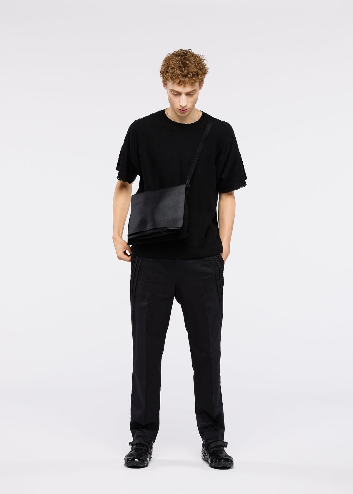 A young man with curly hair stands against a plain background, looking down with one hand in his pocket, wearing the ROLL shoulder bag by GOOD GOODS ISSEY MIYAKE along with an all-black outfit. view 7 of 7