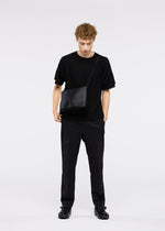 A young man with curly hair stands against a plain background, looking down with one hand in his pocket, wearing the ROLL shoulder bag by GOOD GOODS ISSEY MIYAKE along with an all-black outfit.