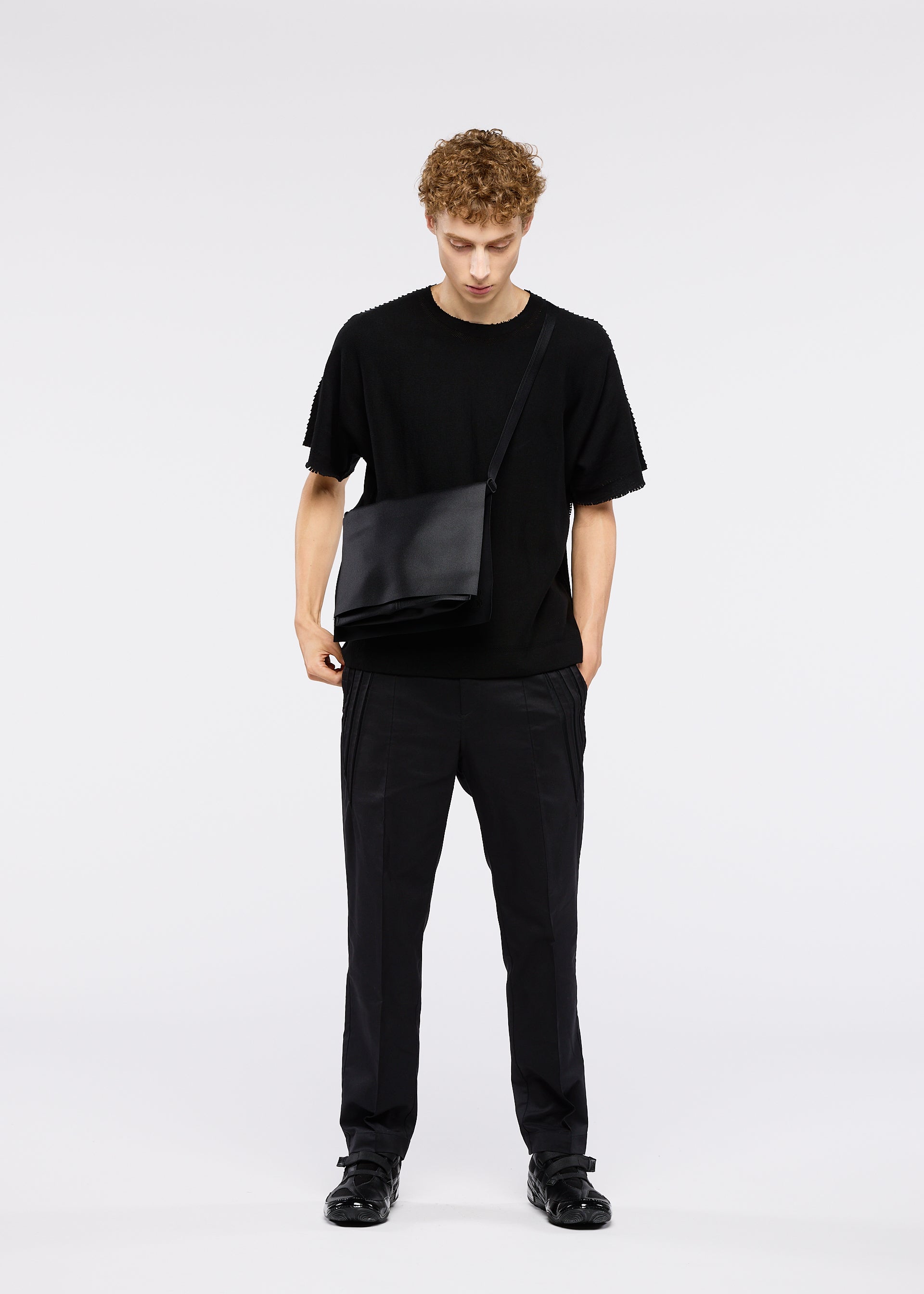 Zoomed view A young man with curly hair stands against a plain background, looking down with one hand in his pocket, wearing the ROLL shoulder bag by GOOD GOODS ISSEY MIYAKE along with an all-black outfit.