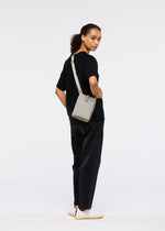 A person with curly hair pulled back stands sideways against a plain white background, wearing a black t-shirt, dark pants, white shoes, and carrying the JABARA shoulder bag by GOOD GOODS ISSEY MIYAKE.