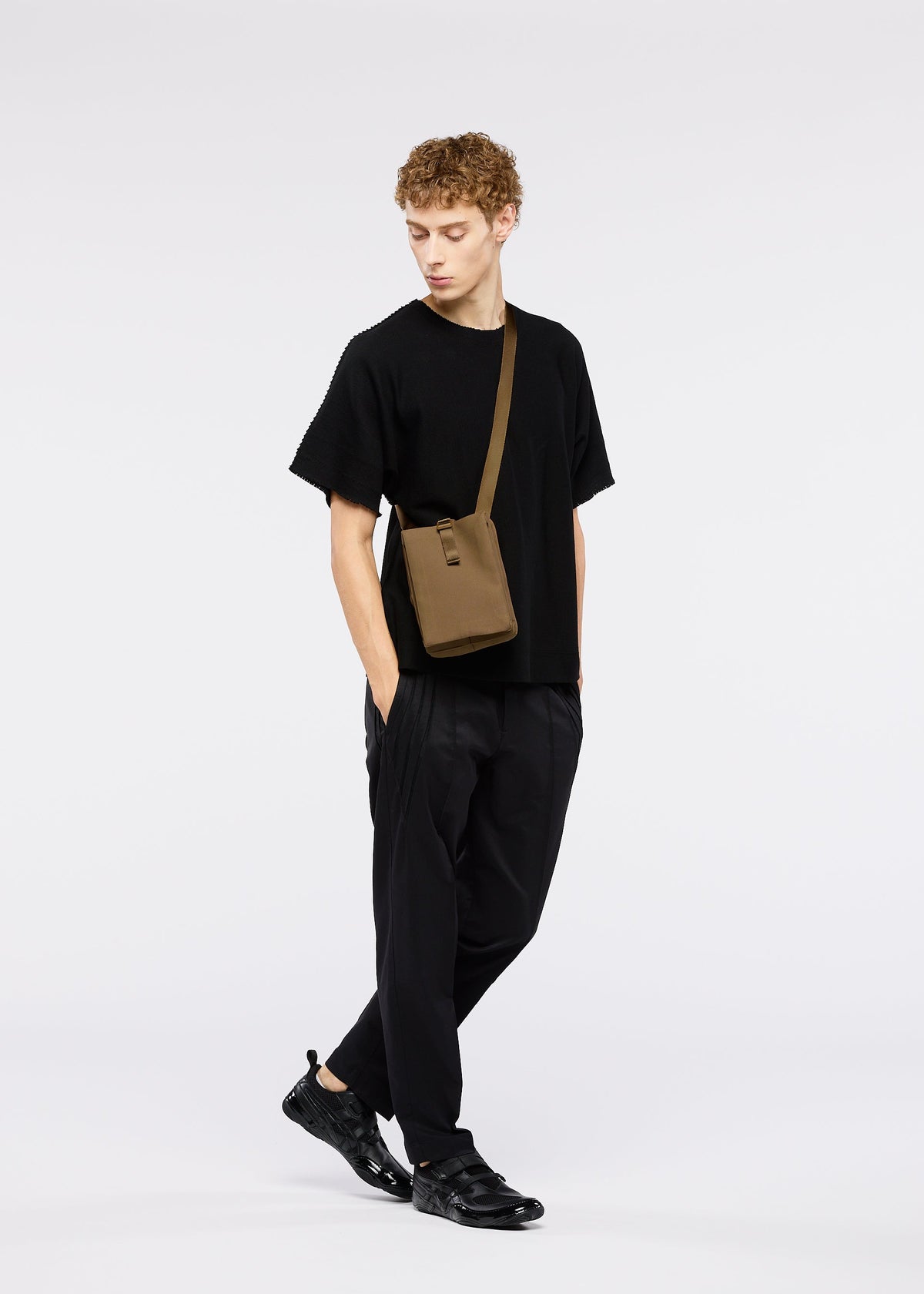 A young person with curly hair wears black clothes and sneakers, carrying the JABARA shoulder bag by GOOD GOODS ISSEY MIYAKE. They stand against a white background, looking down to the side with hands in pockets. view 9 of 9