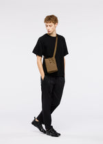 A young person with curly hair wears black clothes and sneakers, carrying the JABARA shoulder bag by GOOD GOODS ISSEY MIYAKE. They stand against a white background, looking down to the side with hands in pockets.