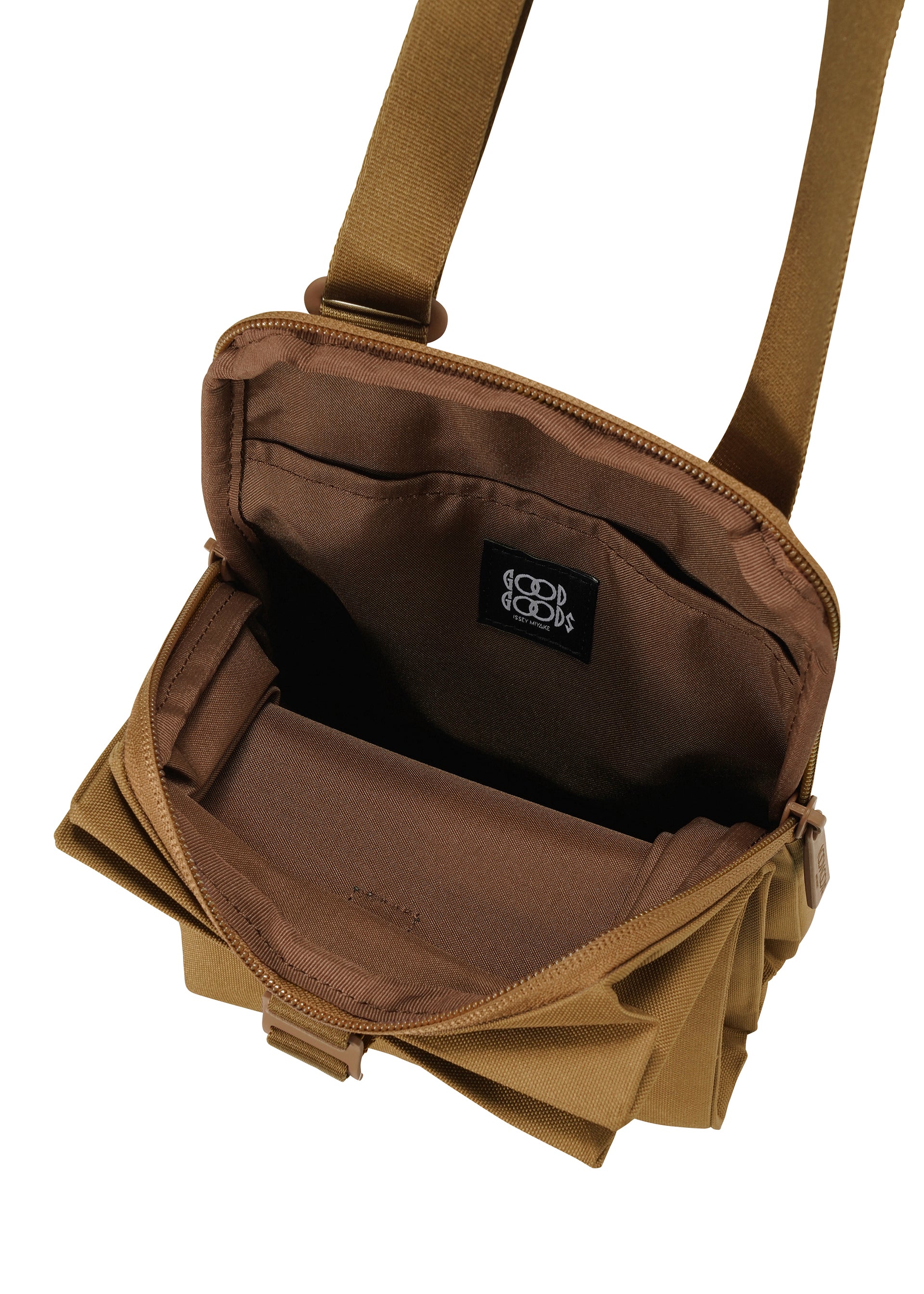 Zoomed view The JABARA by GOOD GOODS ISSEY MIYAKE is a brown pleated gusset bag with a zippered main compartment, brown interior, and a black label with white symbols inside. An adjustable shoulder strap is visible at the top.