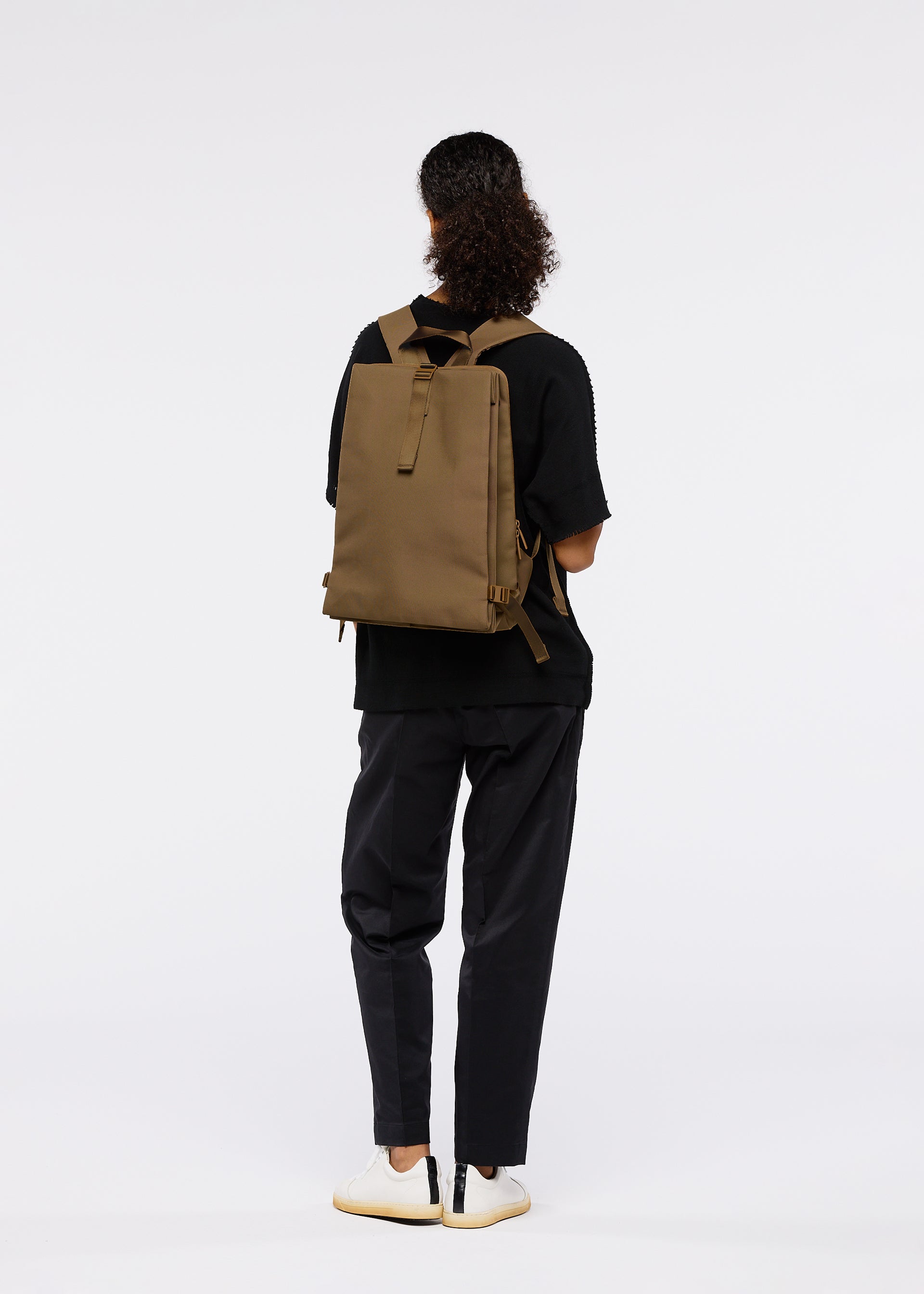 Zoomed view A person with curly hair, wearing a black shirt, dark pants, and white sneakers, stands facing away on a plain white background with the JABARA accordion pleat backpack by GOOD GOODS ISSEY MIYAKE featuring a laptop sleeve.
