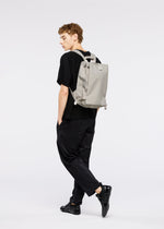 A young man with curly hair, dressed in black, poses sideways with the JABARA backpack by GOOD GOODS ISSEY MIYAKE—featuring accordion pleats and a laptop sleeve—against a plain white background.