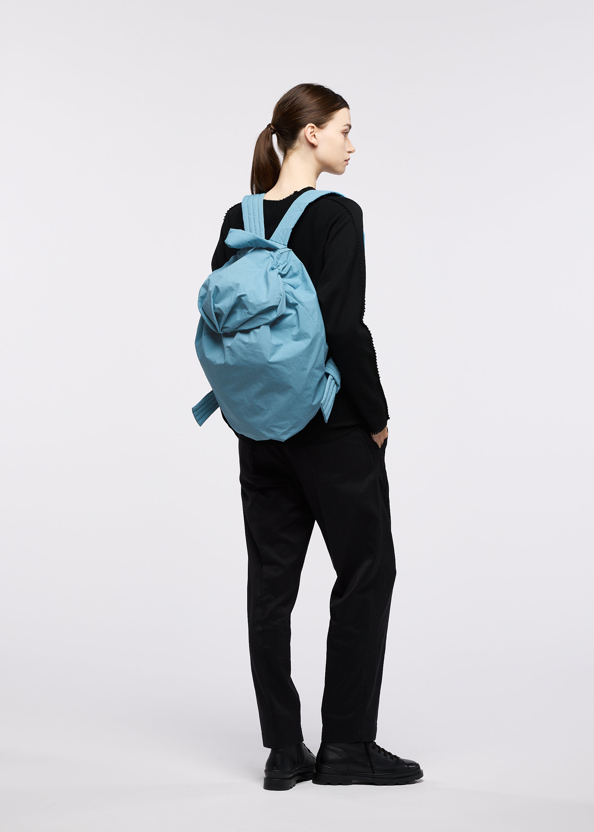 Zoomed view A person stands sideways, facing left, dressed in black attire evoking traditional Japanese workwear and black shoes, carrying the large light blue FUWA FUWA soft backpack by GOOD GOODS ISSEY MIYAKE on their back, set against a plain white background.