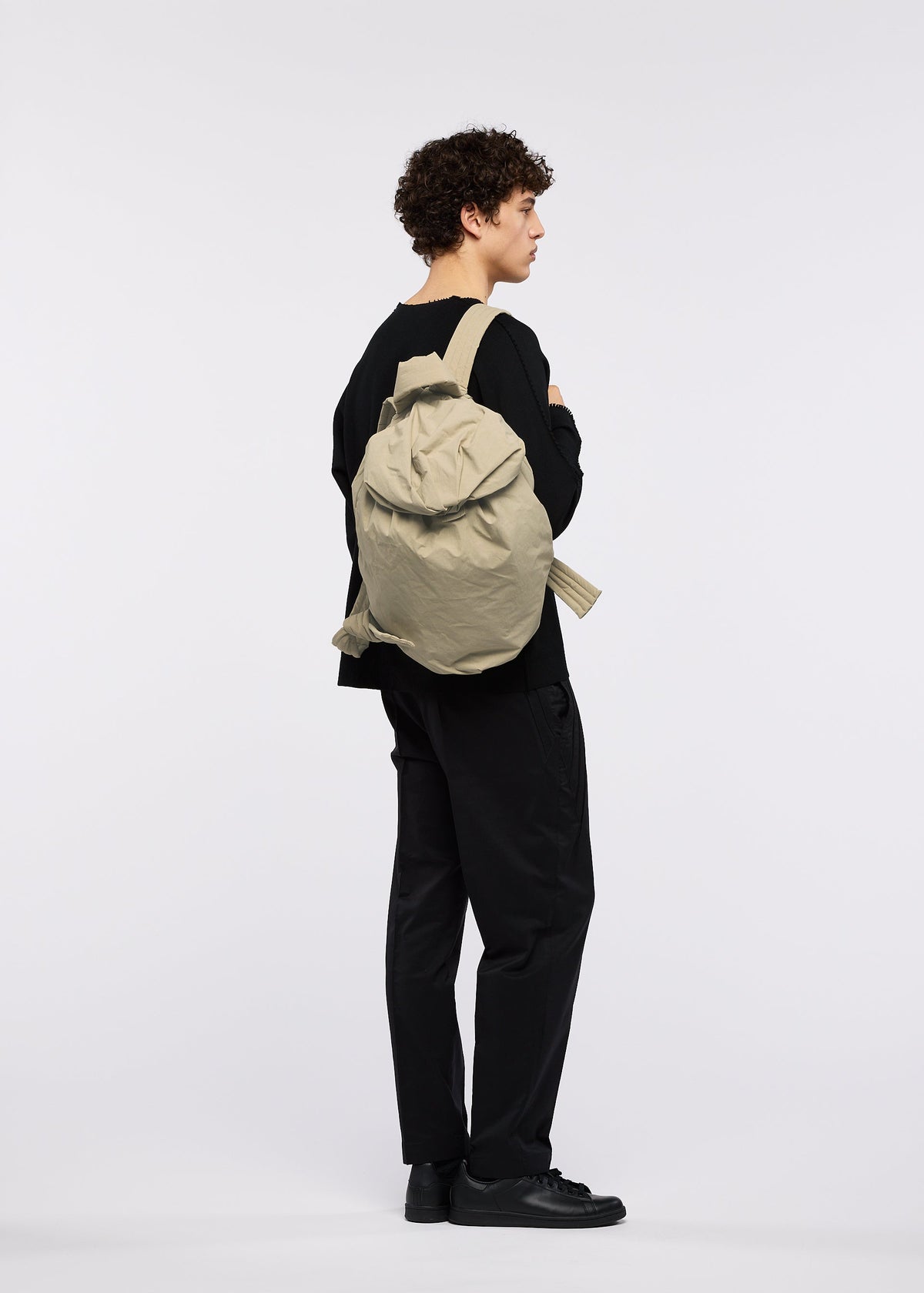 A person with curly hair, dressed entirely in black attire inspired by traditional Japanese workwear and black shoes, stands sideways with arms crossed, carrying the large beige FUWA FUWA soft backpack by GOOD GOODS ISSEY MIYAKE against a plain white background. view 8 of 8