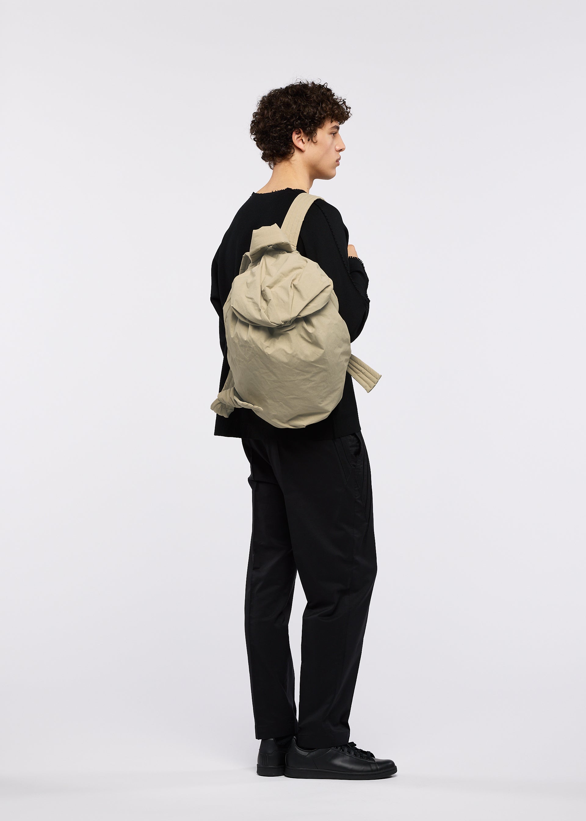 Zoomed view A person with curly hair, dressed entirely in black attire inspired by traditional Japanese workwear and black shoes, stands sideways with arms crossed, carrying the large beige FUWA FUWA soft backpack by GOOD GOODS ISSEY MIYAKE against a plain white background.
