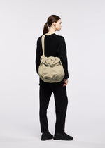 A person with brown hair in a ponytail, dressed in a black outfit and black shoes, stands sideways against a plain white background, carrying the FUWA FUWA shoulder bag by GOOD GOODS ISSEY MIYAKE—a large beige accessory whose simple, elegant design subtly reflects traditional Japanese aesthetics.