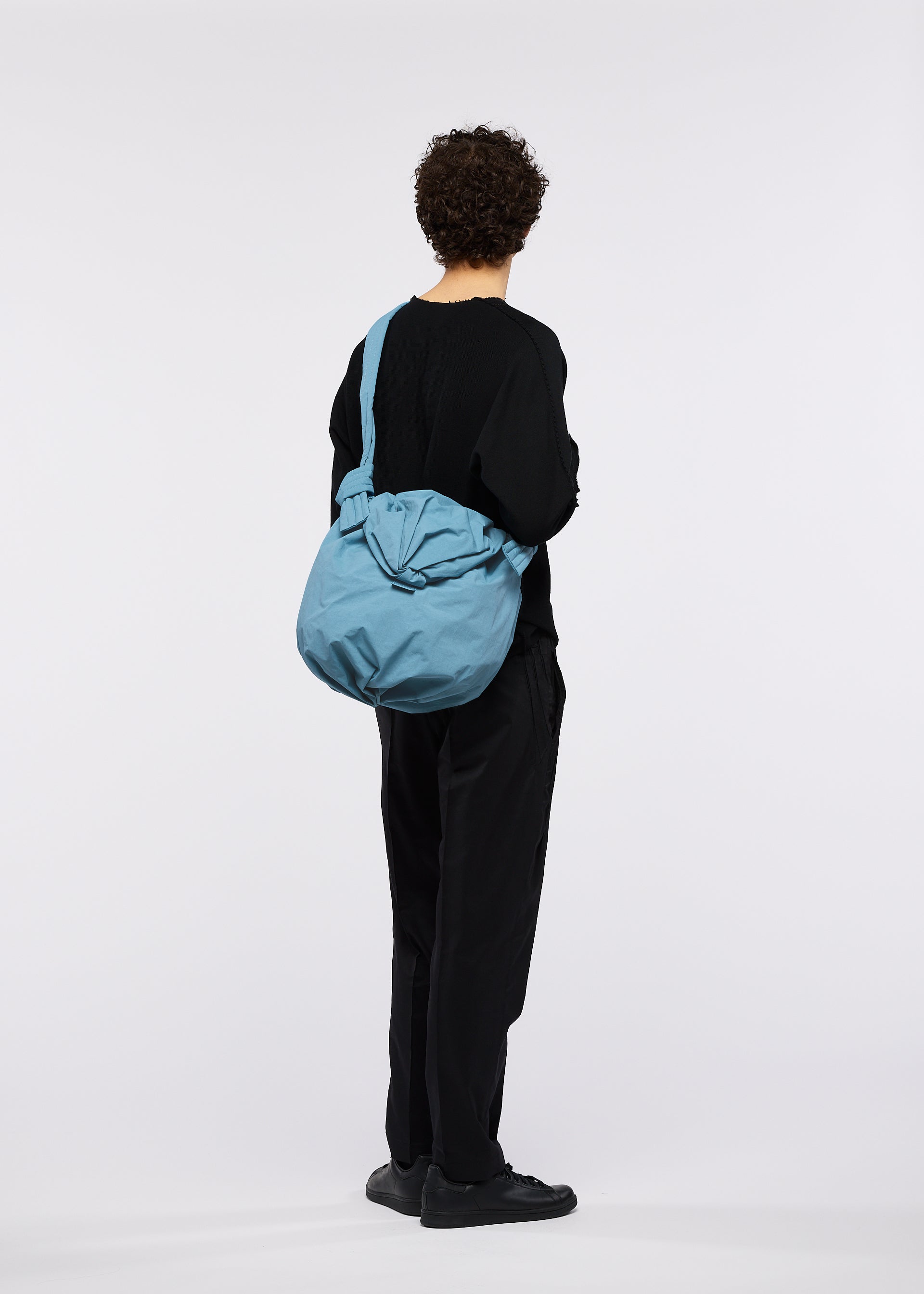 Zoomed view A person with curly hair, dressed in black clothing and shoes, stands facing away against a plain white background, carrying the FUWA FUWA shoulder bag by GOOD GOODS ISSEY MIYAKE—a large, light blue accessory featuring traditional Japanese zero-waste design elements.