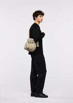 A person with curly hair stands sideways against a plain white background, wearing the FUWA FUWA black outfit by GOOD GOODS ISSEY MIYAKE, paired with black shoes and carrying a beige shoulder bag over their shoulder.
