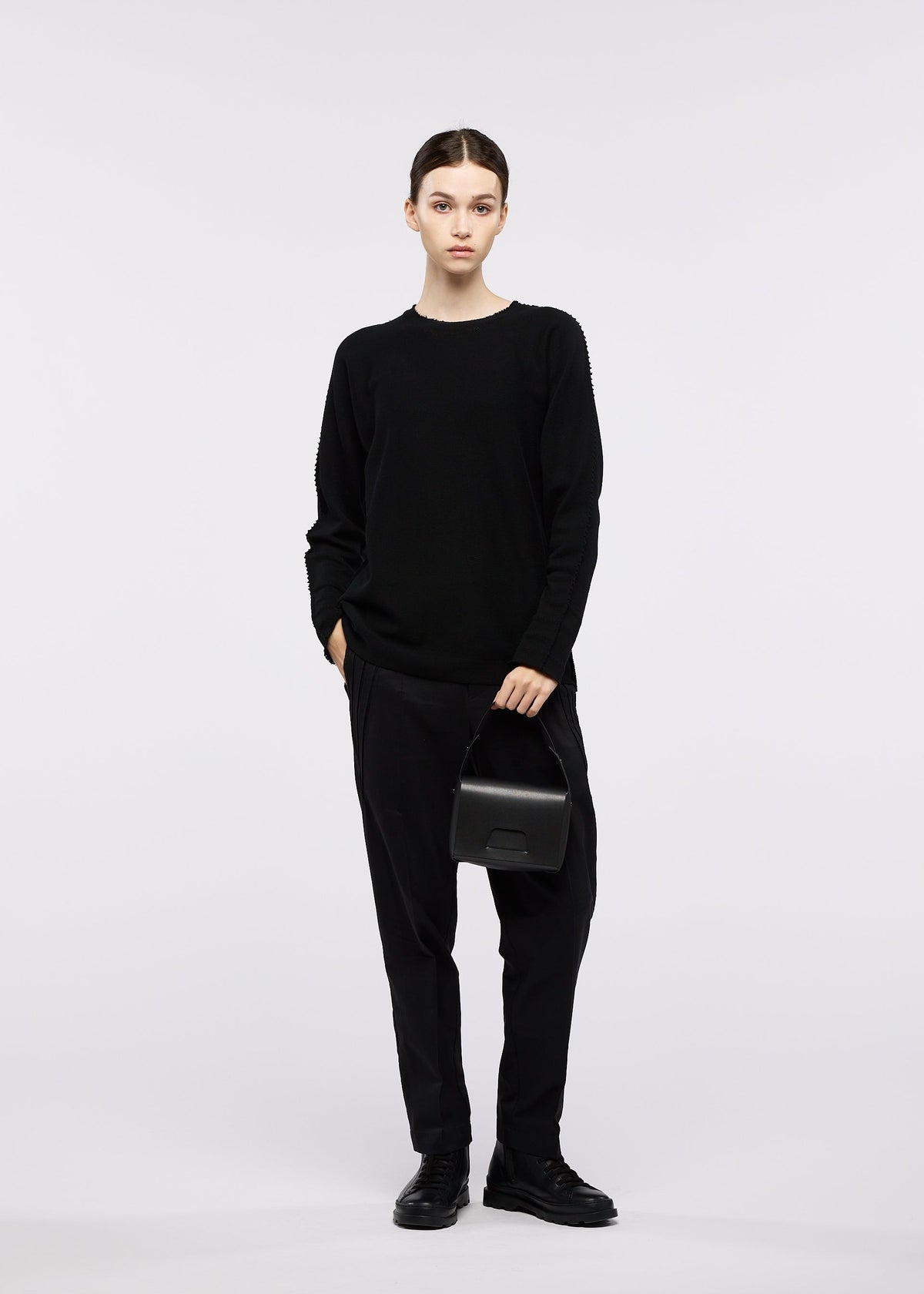 A person stands against a plain background, wearing a black sweater, black pants, and black shoes, holding the GOOD GOODS ISSEY MIYAKE BOX bag and looking at the camera with a neutral expression. view 2 of 4
