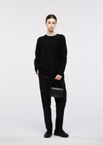 A person stands against a plain background, wearing a black sweater, black pants, and black shoes, holding the GOOD GOODS ISSEY MIYAKE BOX bag and looking at the camera with a neutral expression.