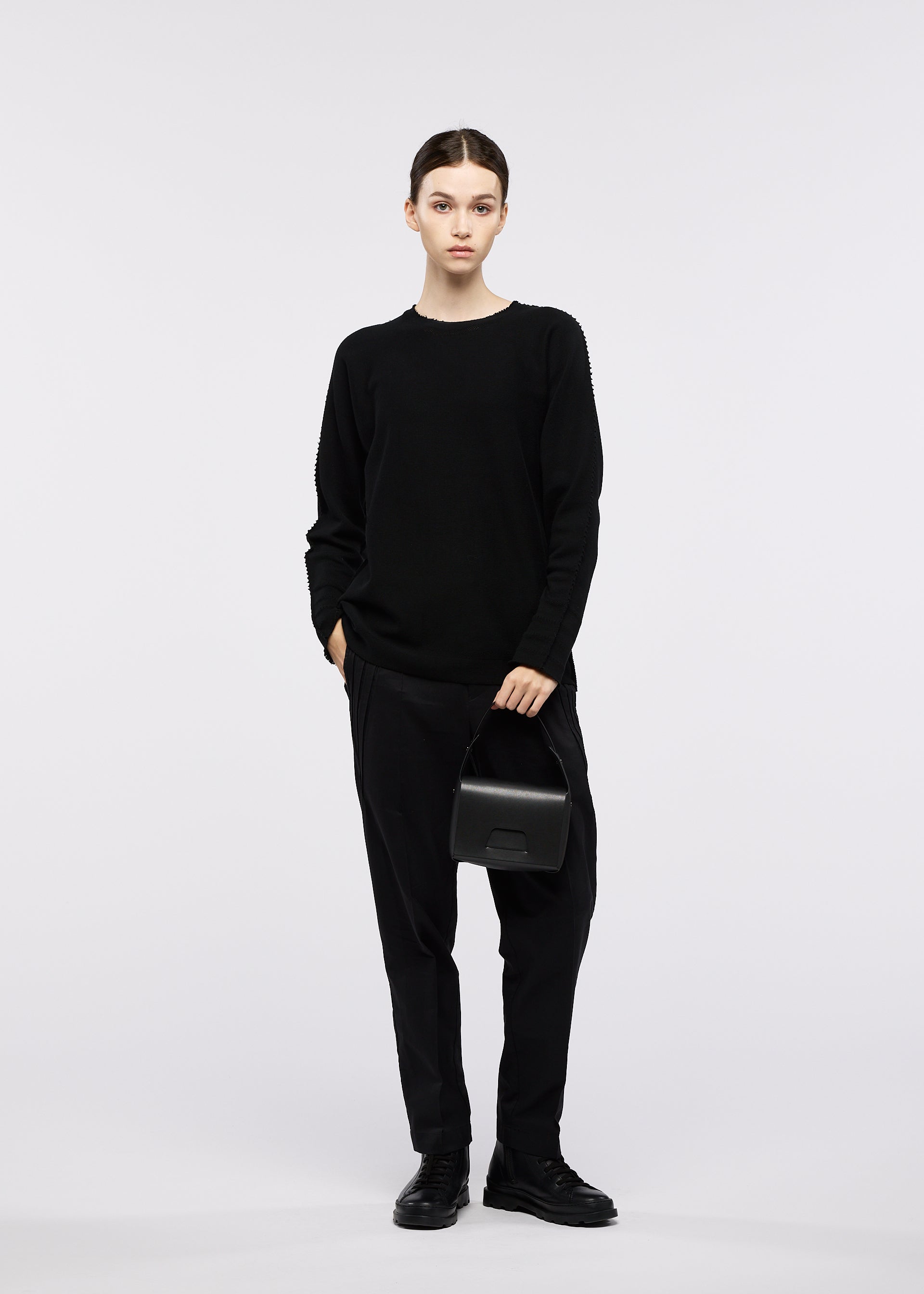 Zoomed view A person stands against a plain background, wearing a black sweater, black pants, and black shoes, holding the GOOD GOODS ISSEY MIYAKE BOX bag and looking at the camera with a neutral expression.