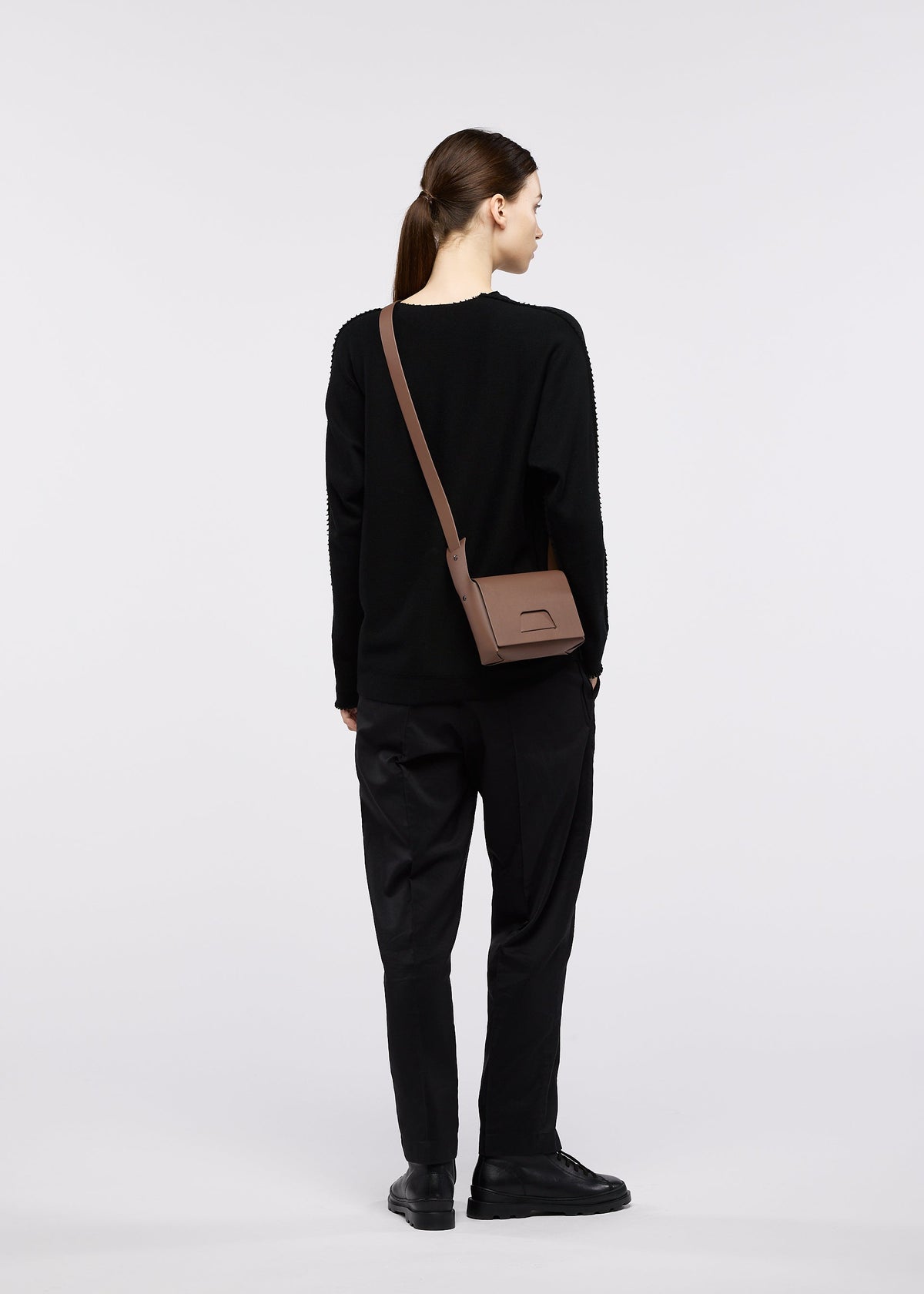 A person with brown hair in a ponytail, dressed in a black sweater, black pants, and black shoes, stands facing away while carrying the BOX shoulder bag by GOOD GOODS ISSEY MIYAKE against a plain light-colored background. view 3 of 4