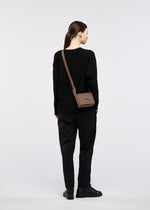 A person with brown hair in a ponytail, dressed in a black sweater, black pants, and black shoes, stands facing away while carrying the BOX shoulder bag by GOOD GOODS ISSEY MIYAKE against a plain light-colored background.