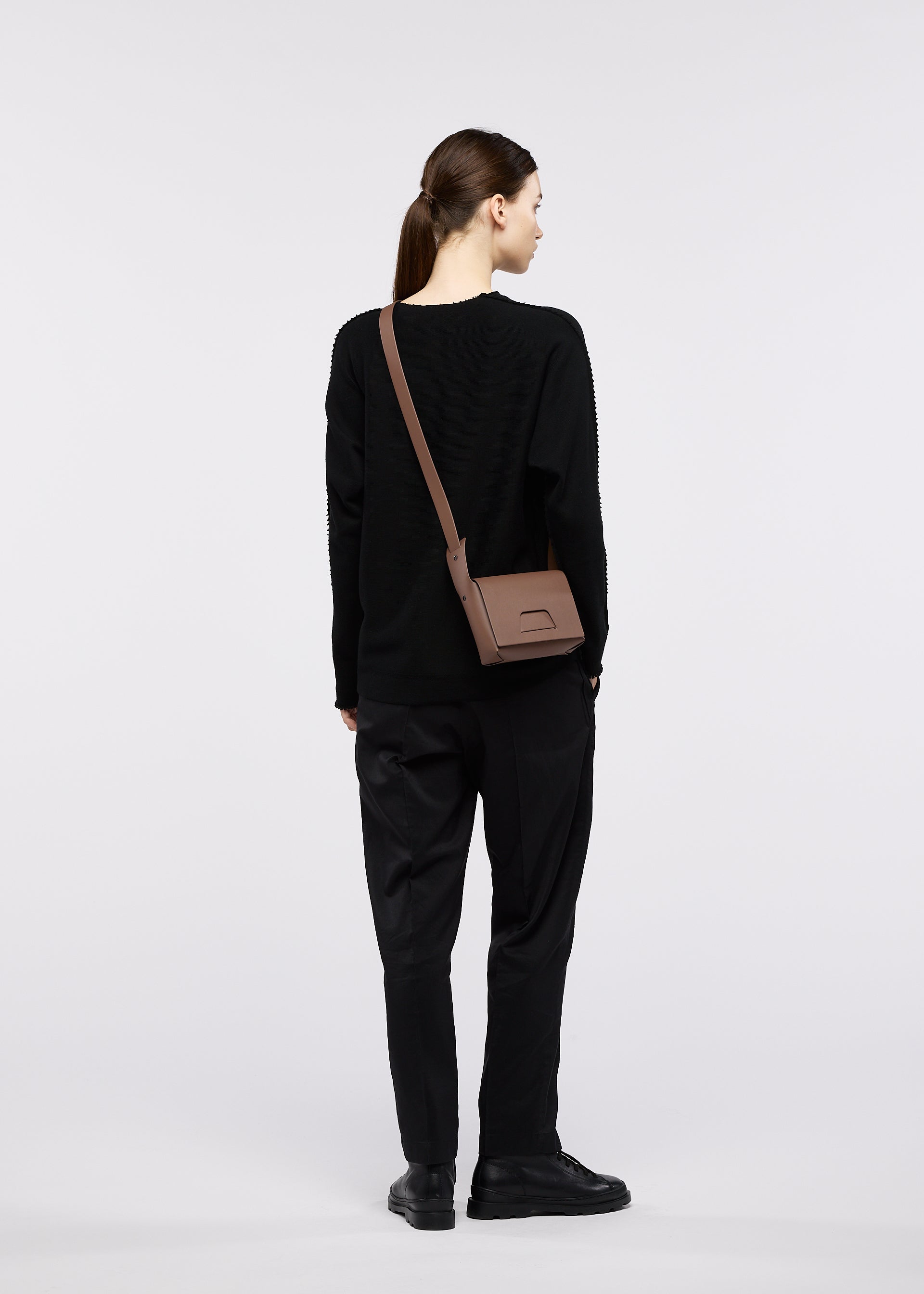 Zoomed view A person with brown hair in a ponytail, dressed in a black sweater, black pants, and black shoes, stands facing away while carrying the BOX shoulder bag by GOOD GOODS ISSEY MIYAKE against a plain light-colored background.