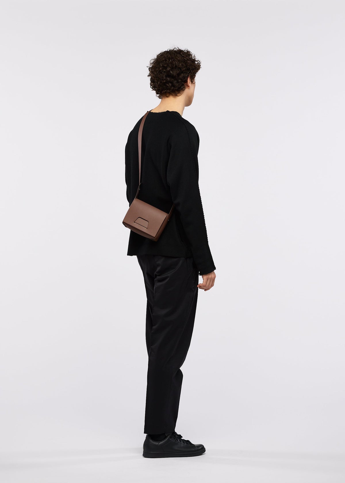 A person with curly hair, dressed entirely in black, stands facing away against a plain white background. They are wearing the BOX bag by GOOD GOODS ISSEY MIYAKE slung across their shoulder, paired with a black long-sleeve top, black pants, and black shoes. view 4 of 4