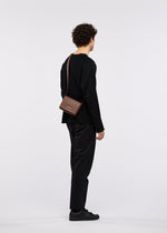 A person with curly hair, dressed entirely in black, stands facing away against a plain white background. They are wearing the BOX bag by GOOD GOODS ISSEY MIYAKE slung across their shoulder, paired with a black long-sleeve top, black pants, and black shoes.