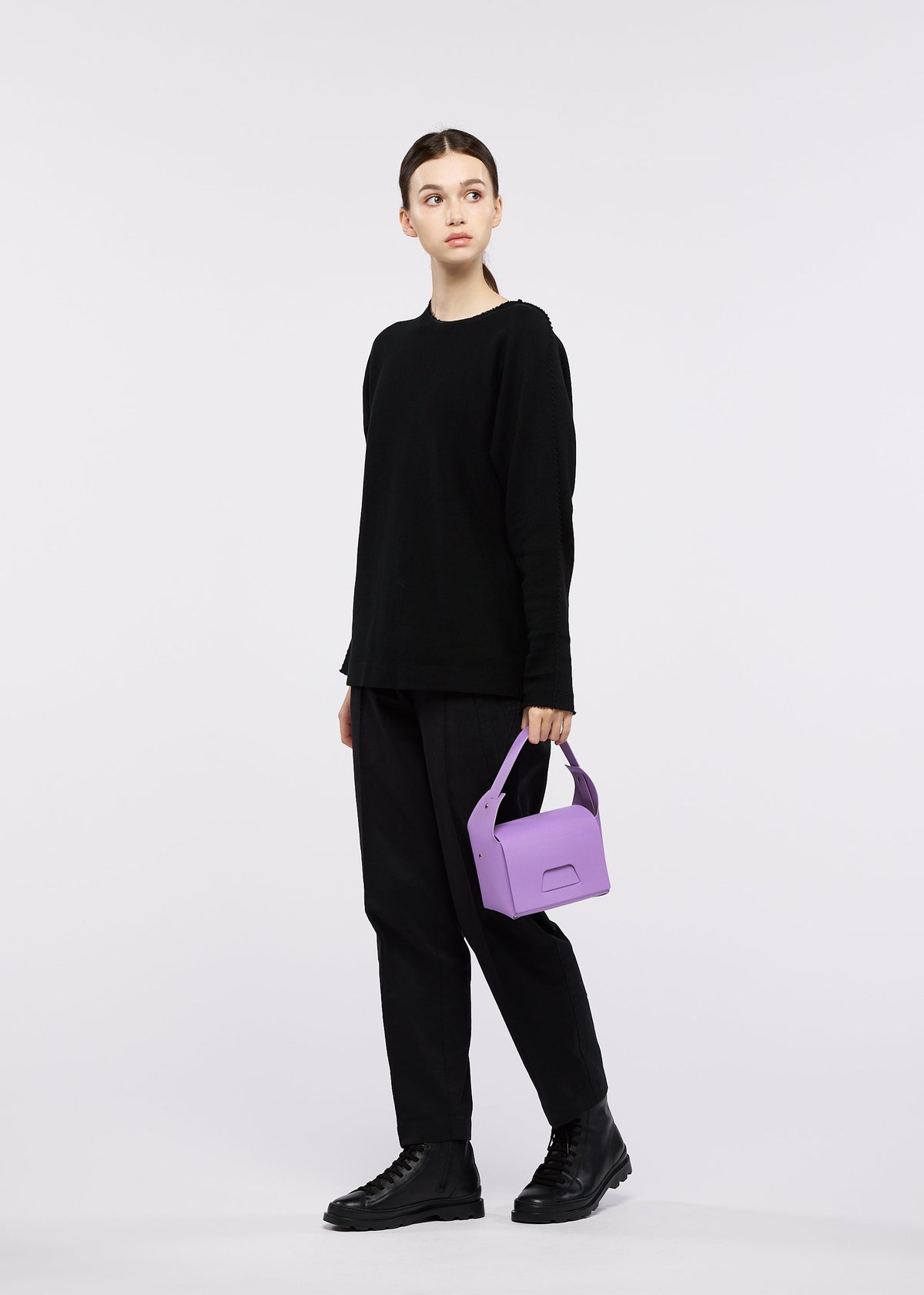 A young woman dressed in a black outfit and boots stands against a white background, holding the GOOD GOODS ISSEY MIYAKE BOX bag in purple and gazing slightly to the side. view 2 of 2