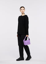 A young woman dressed in a black outfit and boots stands against a white background, holding the GOOD GOODS ISSEY MIYAKE BOX bag in purple and gazing slightly to the side.