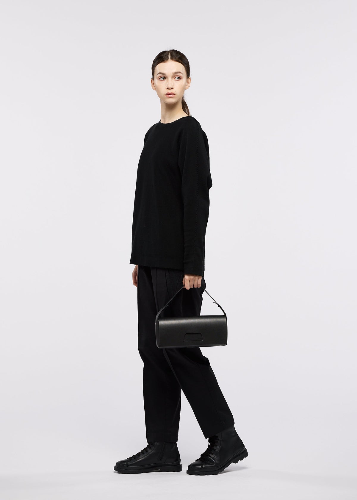 A person stands against a white background wearing a black long-sleeve shirt, black pants, and black shoes, holding the BOX bag by GOOD GOODS ISSEY MIYAKE. They are looking slightly to the left. view 7 of 8