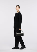 A person stands against a white background wearing a black long-sleeve shirt, black pants, and black shoes, holding the BOX bag by GOOD GOODS ISSEY MIYAKE. They are looking slightly to the left.