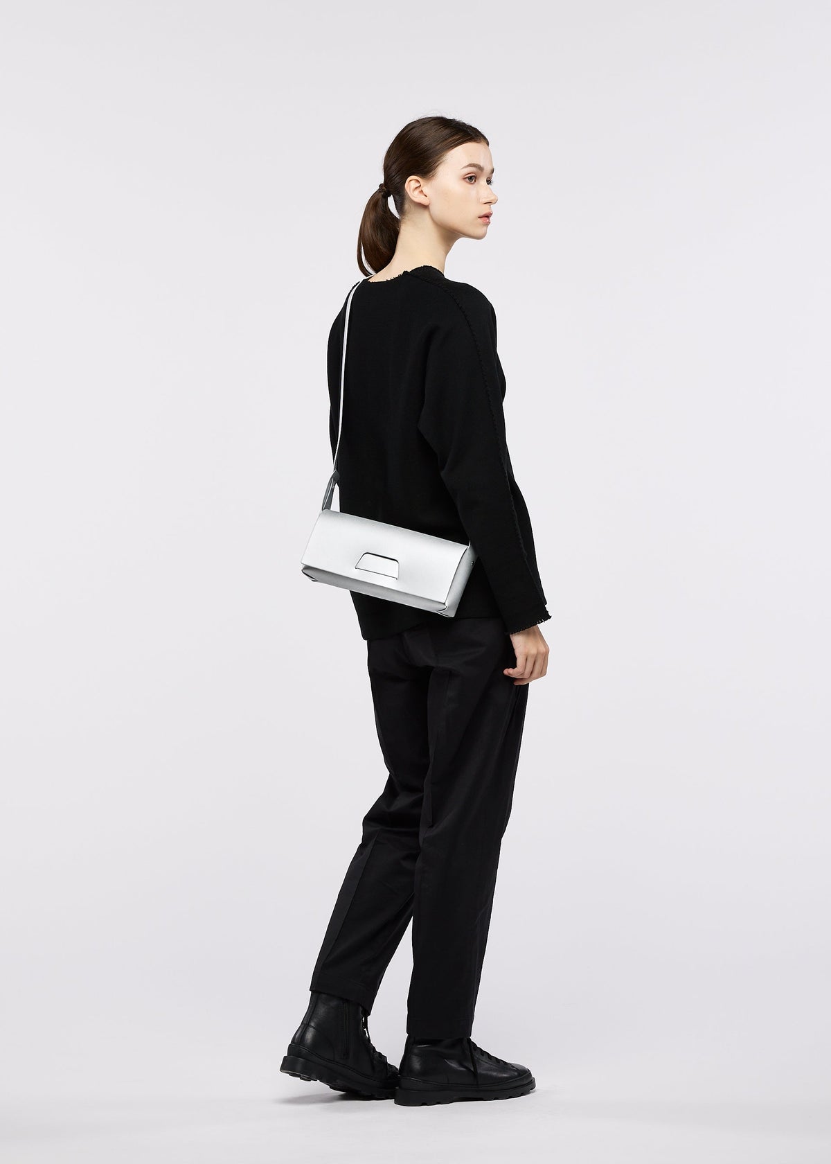 A woman stands in profile wearing a black outfit with matching black shoes, carrying the BOX shoulder bag by GOOD GOODS ISSEY MIYAKE in white. Her brown hair is tied back, and she poses against a plain, light-colored background. view 8 of 8