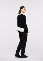A woman stands in profile wearing a black outfit with matching black shoes, carrying the BOX shoulder bag by GOOD GOODS ISSEY MIYAKE in white. Her brown hair is tied back, and she poses against a plain, light-colored background.