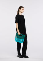 A person stands against a plain white background, dressed in all black and holding the translucent teal MOKKO BI-COLOR tote bag by GOOD GOODS ISSEY MIYAKE at their side.