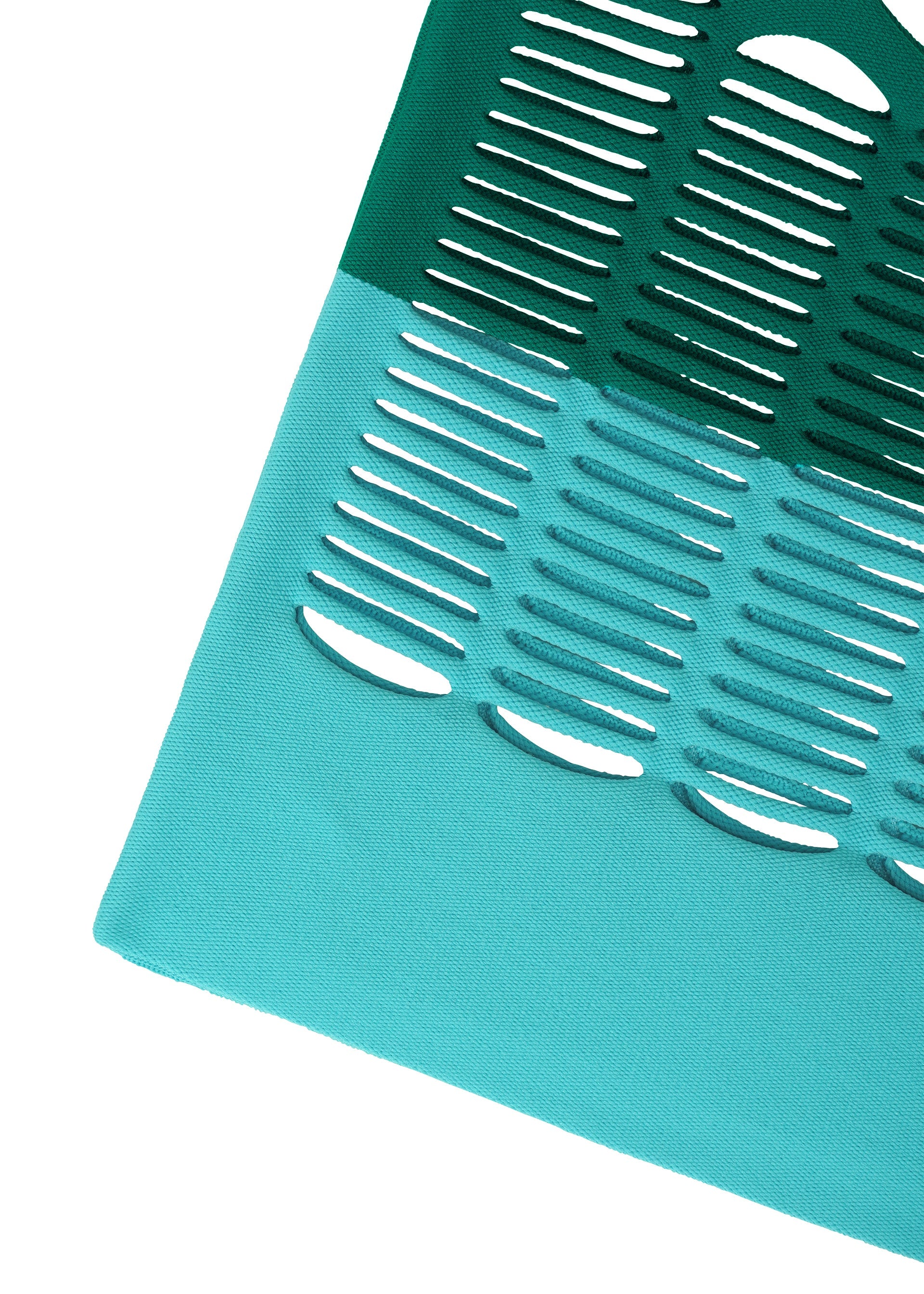 Zoomed view A close-up of the MOKKO BI-COLOR chair seat by GOOD GOODS ISSEY MIYAKE shows two teal shades and oval cutout patterns, recalling a stylish knit bag. The seat's corner appears against a white background.
