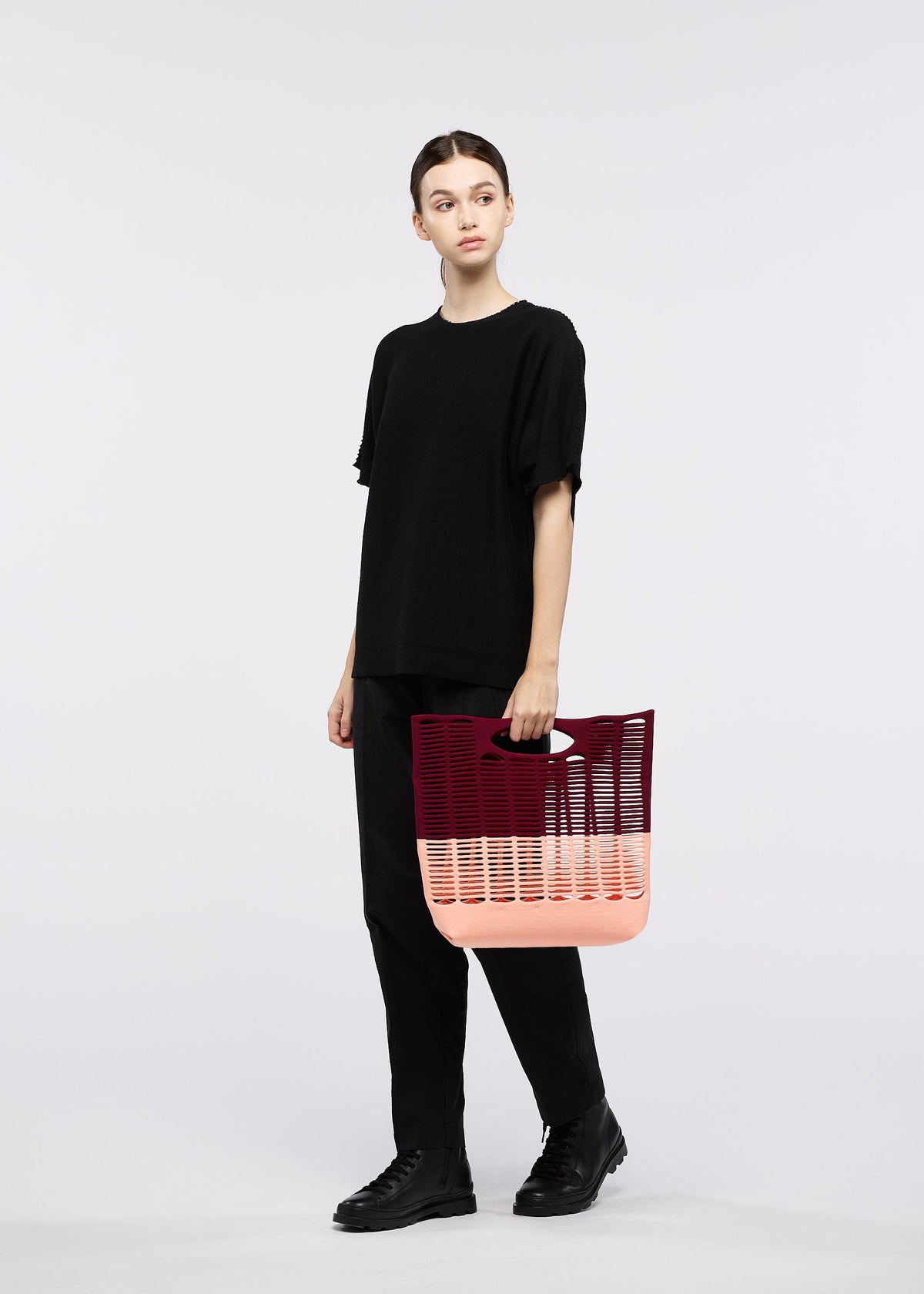 A person dressed in black stands against a white background, holding the GOOD GOODS ISSEY MIYAKE MOKKO BI-COLOR tote bag in pink and burgundy knit. view 8 of 9