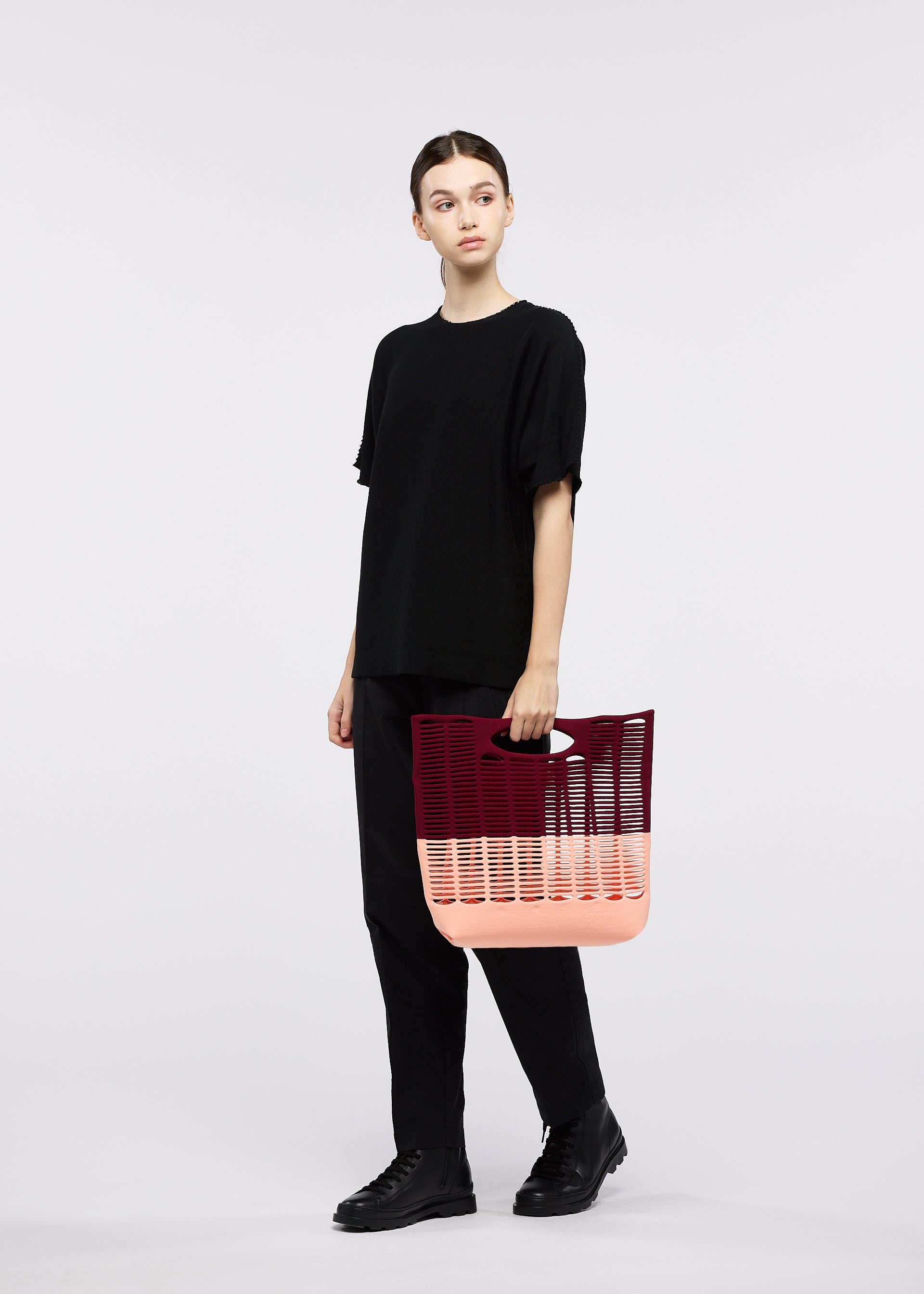 Zoomed view A person dressed in black stands against a white background, holding the GOOD GOODS ISSEY MIYAKE MOKKO BI-COLOR tote bag in pink and burgundy knit.