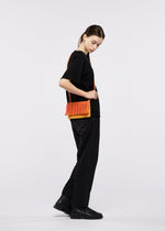 A young woman in black clothing and boots stands sideways against a plain white background, wearing the orange MOKKO BI-COLOR tote bag by GOOD GOODS ISSEY MIYAKE.