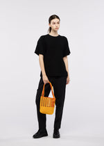 A person dressed in black stands against a plain white background, holding the bright yellow MOKKO BI-COLOR tote bag by GOOD GOODS ISSEY MIYAKE.