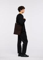 A person with curly hair, dressed in all black, stands in profile against a white background, holding the GOOD GOODS ISSEY MIYAKE MOKKO MONO GRIP—a dark rectangular tote bag featuring a cut-out pattern and an adjustable shoulder strap.