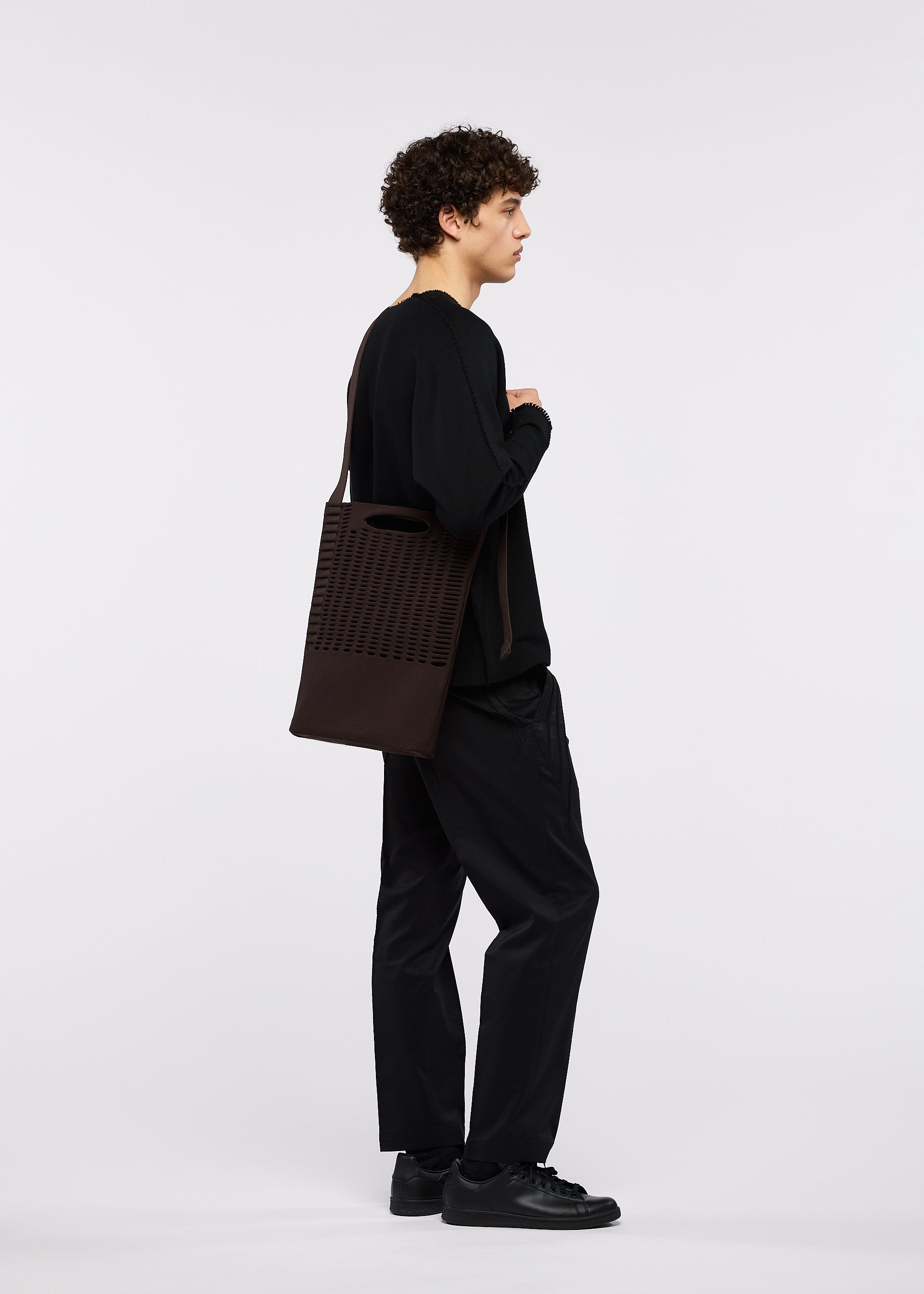 Zoomed view A person with curly hair, dressed in all black, stands in profile against a white background, holding the GOOD GOODS ISSEY MIYAKE MOKKO MONO GRIP—a dark rectangular tote bag featuring a cut-out pattern and an adjustable shoulder strap.