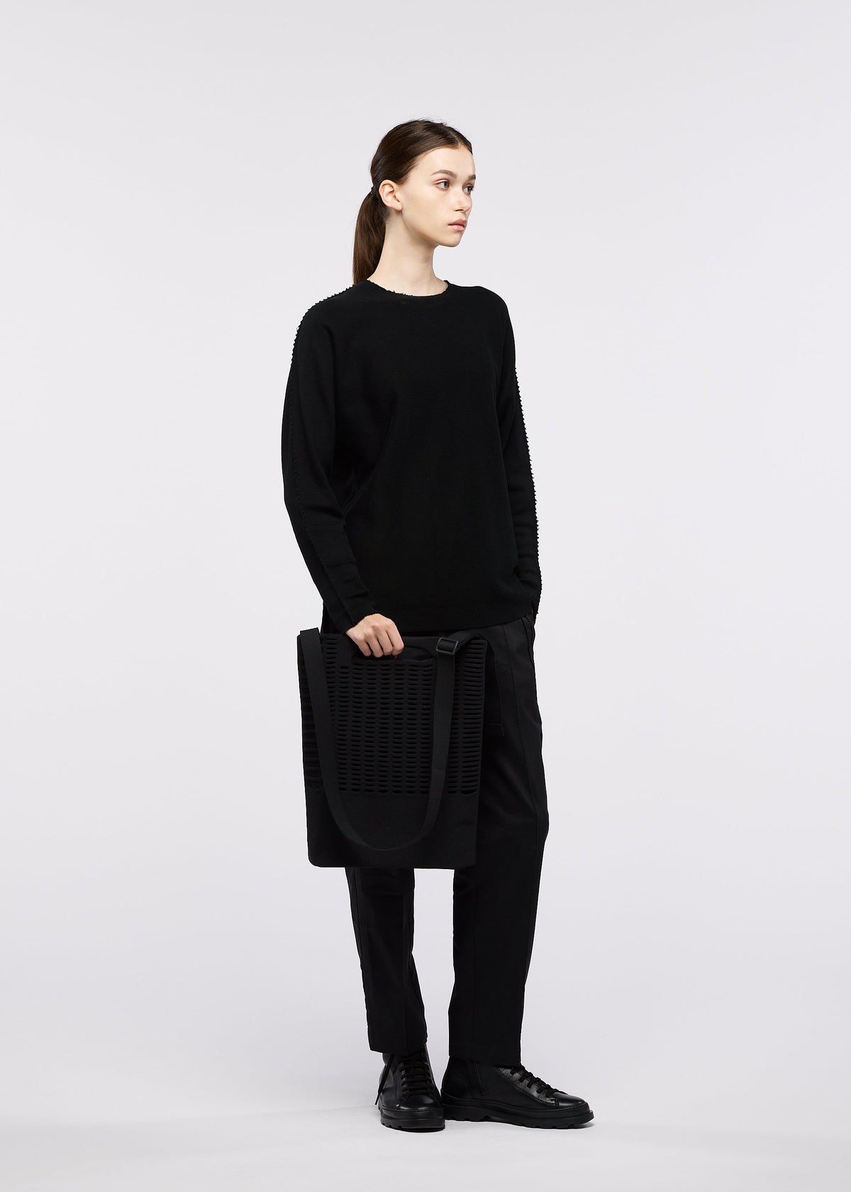 A person dressed in all-black stands holding the MOKKO MONO GRIP business bag by GOOD GOODS ISSEY MIYAKE, featuring an adjustable shoulder strap. The background is plain white, and the individual gazes to the side with a neutral expression. view 6 of 6