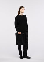 A person dressed in all-black stands holding the MOKKO MONO GRIP business bag by GOOD GOODS ISSEY MIYAKE, featuring an adjustable shoulder strap. The background is plain white, and the individual gazes to the side with a neutral expression.