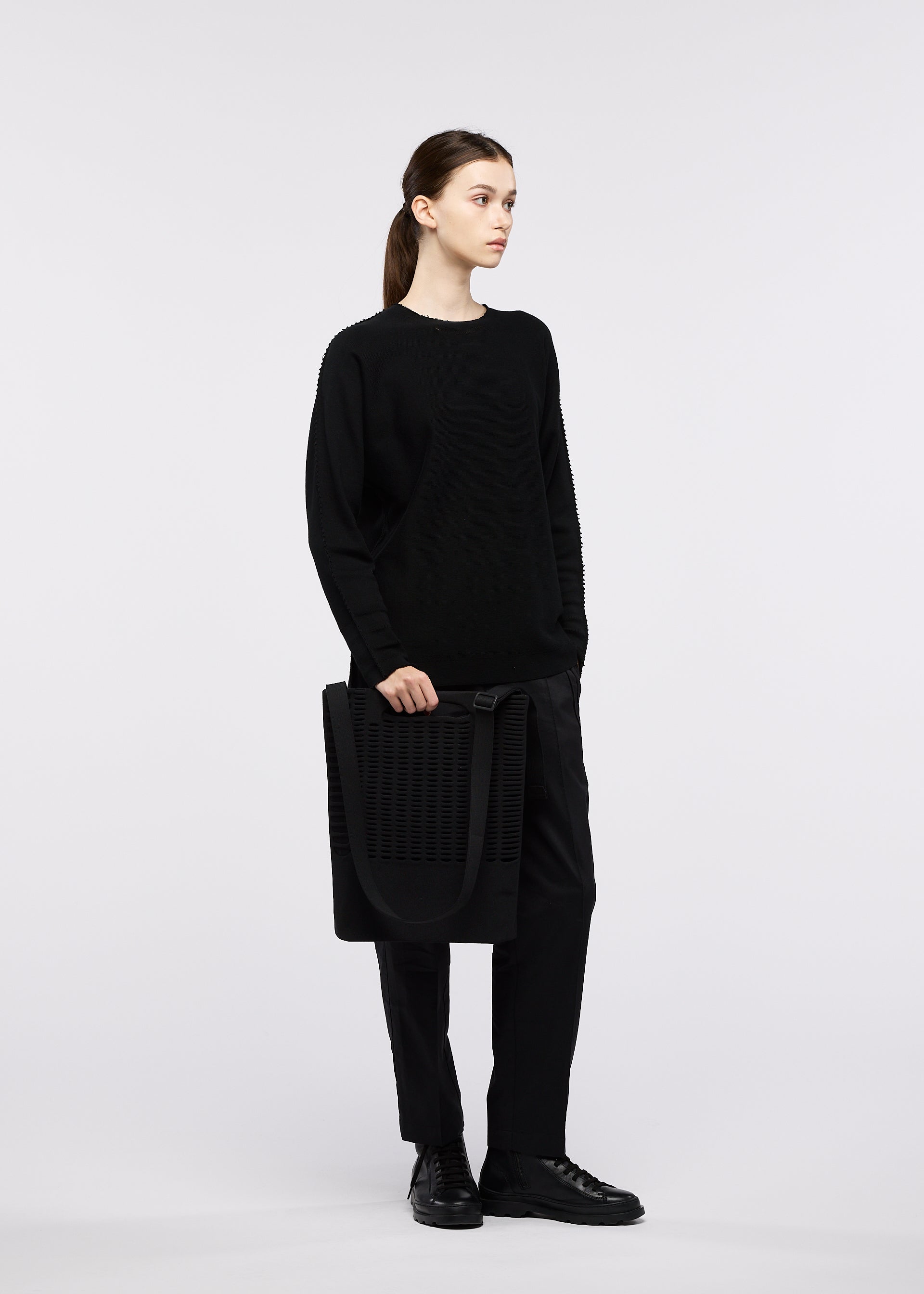 Zoomed view A person dressed in all-black stands holding the MOKKO MONO GRIP business bag by GOOD GOODS ISSEY MIYAKE, featuring an adjustable shoulder strap. The background is plain white, and the individual gazes to the side with a neutral expression.