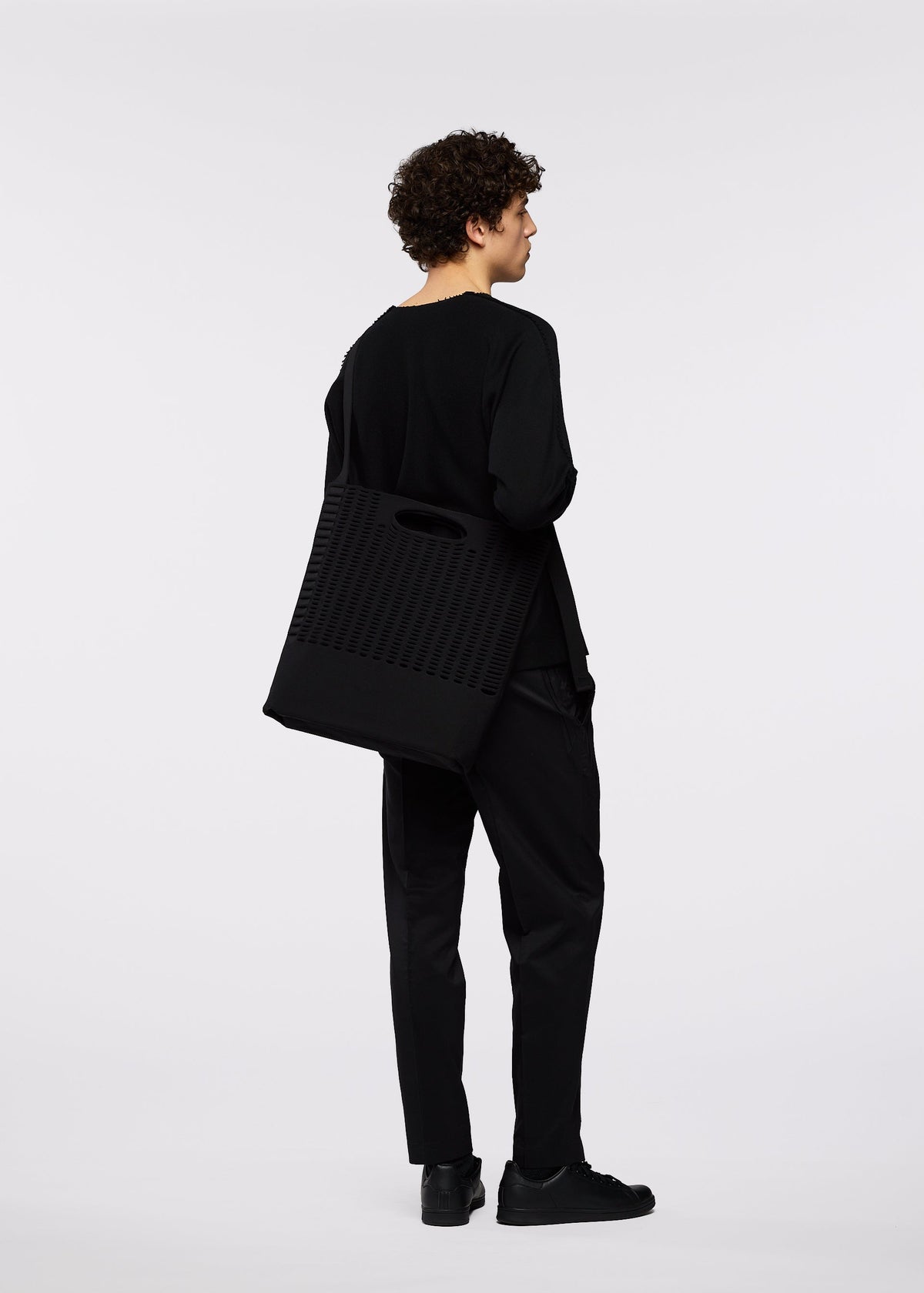 A person with curly hair, dressed in all black, stands facing away, holding a large black business bag with an adjustable shoulder strap over their shoulder against a plain white background. view 5 of 6