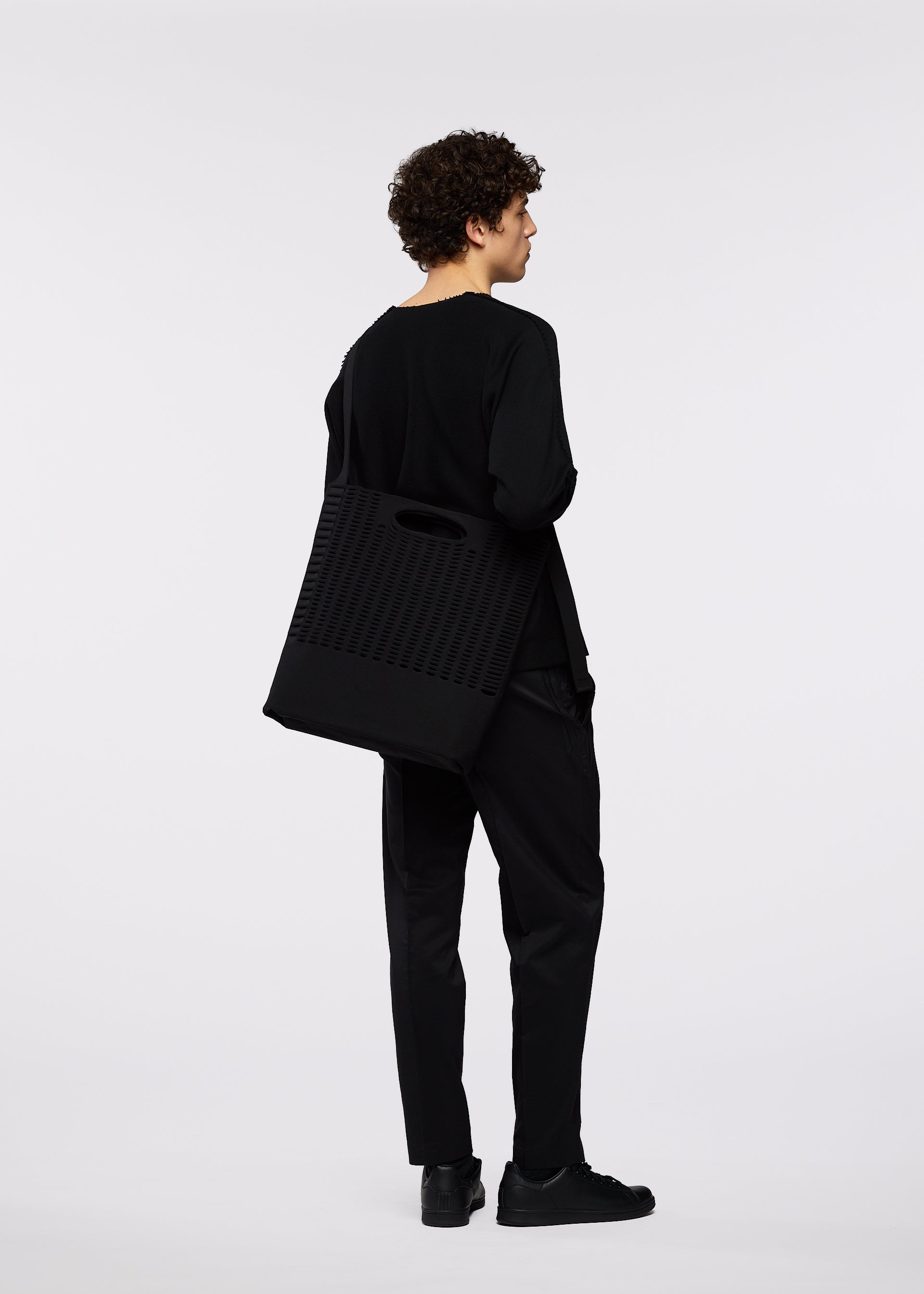 Zoomed view A person with curly hair, dressed in all black, stands facing away, holding a large black business bag with an adjustable shoulder strap over their shoulder against a plain white background.