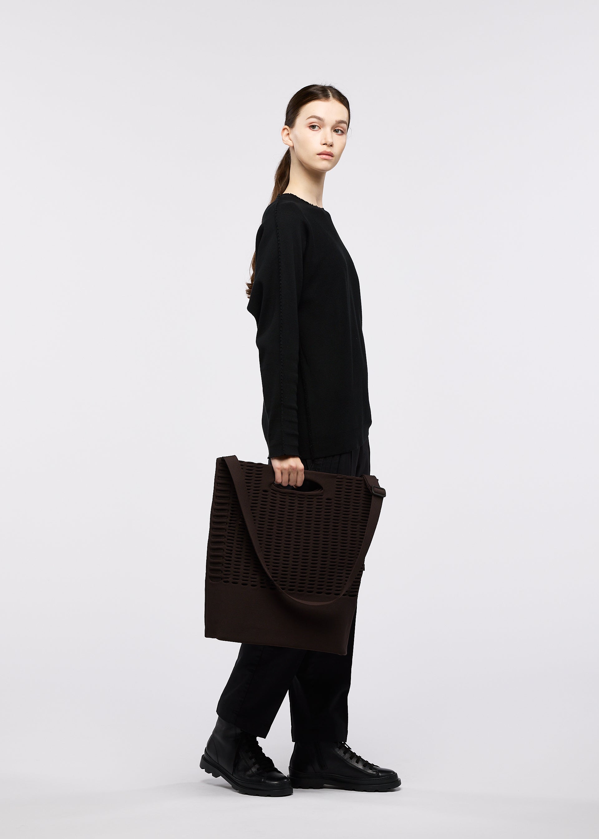Zoomed view A person with long hair in a low ponytail, dressed in black clothing and black shoes, stands in profile holding the MOKKO MONO GRIP tote bag by GOOD GOODS ISSEY MIYAKE—with perforated details and an adjustable shoulder strap—against a plain white background.