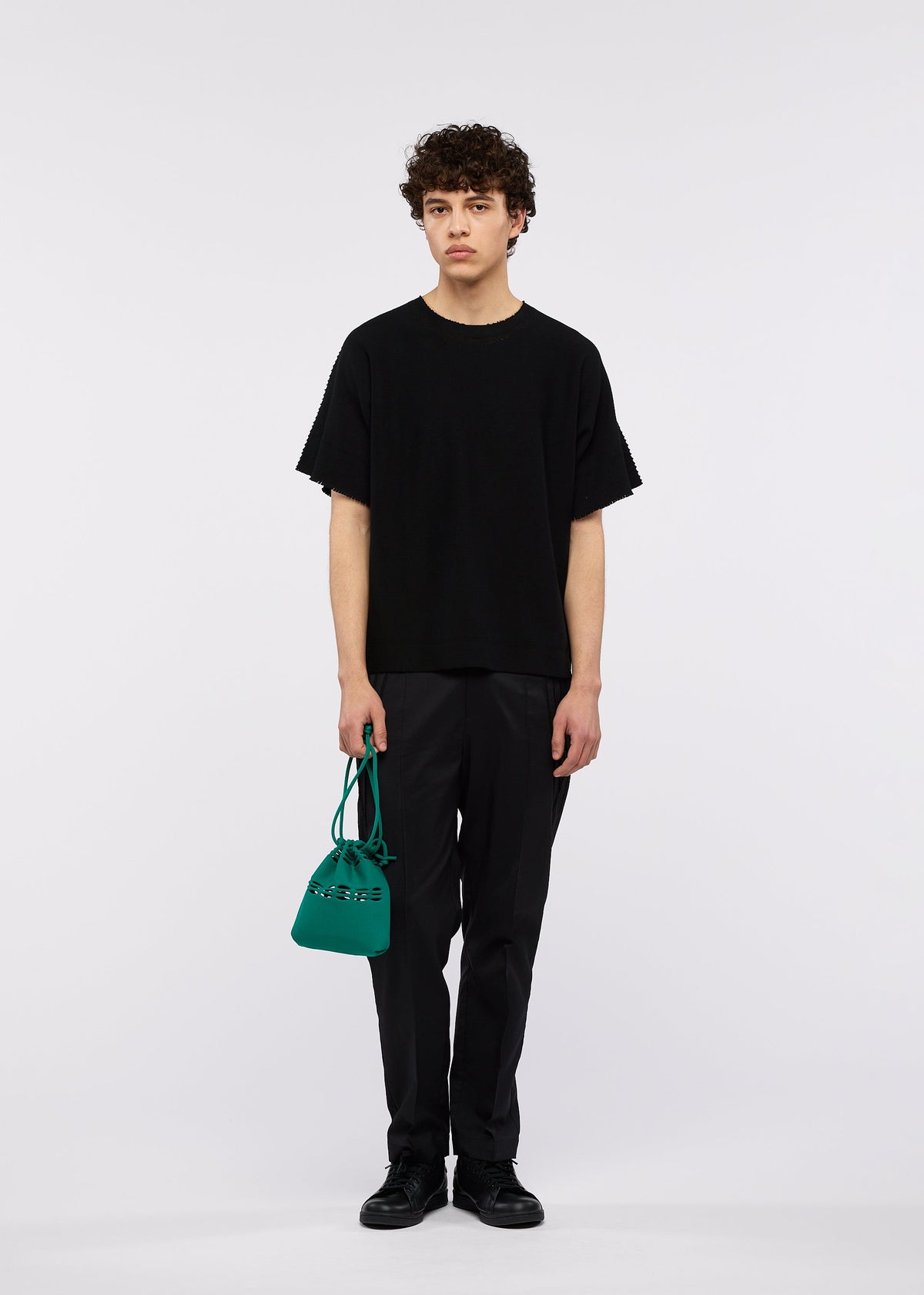A young person with curly hair stands against a white background wearing black clothes and shoes, holding the GOOD GOODS ISSEY MIYAKE MOKKO POCKET BI-COLOR mini bag in green. view 9 of 9
