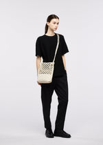A person in all black stands against a plain light background, carrying the cream-colored GOOD GOODS ISSEY MIYAKE MOKKO WASHI crossbody bag crafted from washi paper.