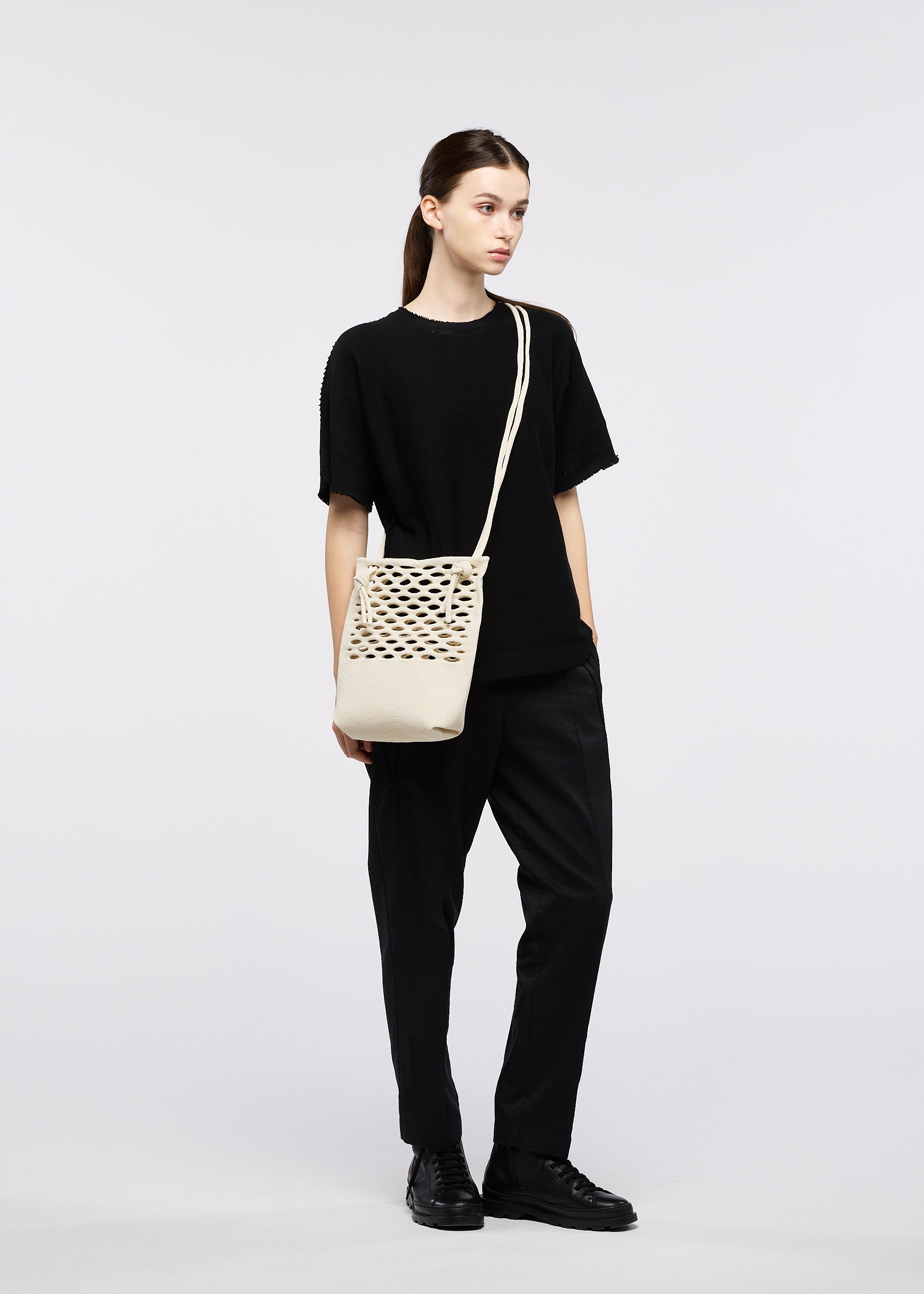 Zoomed view A person in all black stands against a plain light background, carrying the cream-colored GOOD GOODS ISSEY MIYAKE MOKKO WASHI crossbody bag crafted from washi paper.