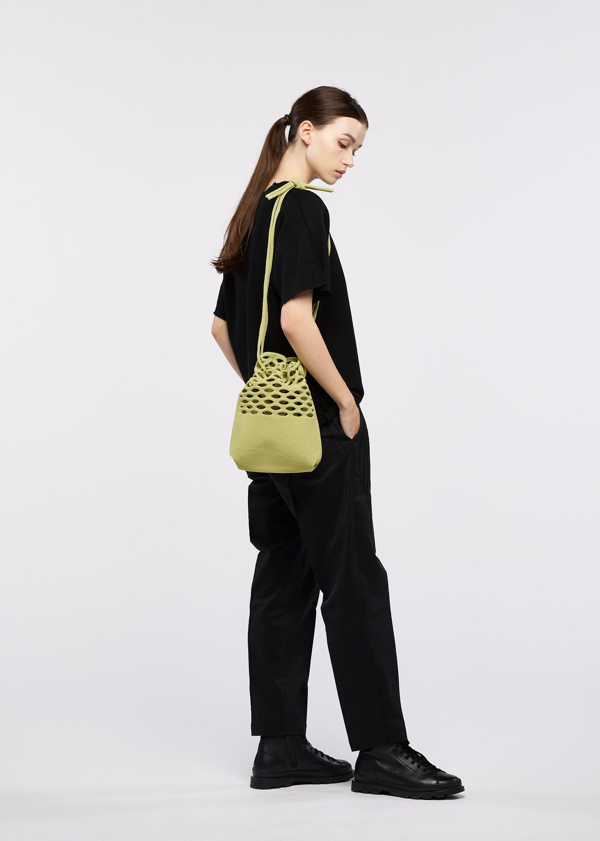 A person with long brown hair in a low ponytail, dressed in black, stands sideways with hands in pockets, carrying a green MOKKO WASHI shoulder bag by GOOD GOODS ISSEY MIYAKE. The background is plain white. view 12 of 13